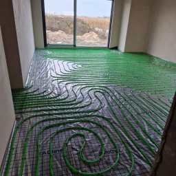 Hydro Gorzów - Zielone rury ogrzewania podłogowego ułożone na folii z siatką w surowym wnętrzu z oknem na zewnątrz. Widoczne elementy mocujące rury.