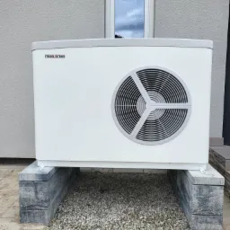 Biała jednostka zewnętrzna pompy ciepła marki Stiebel Eltron, zamontowana na betonowych bloczkach z podsypką żwirową, na tle jasnoszarej elewacji budynku z oknem.