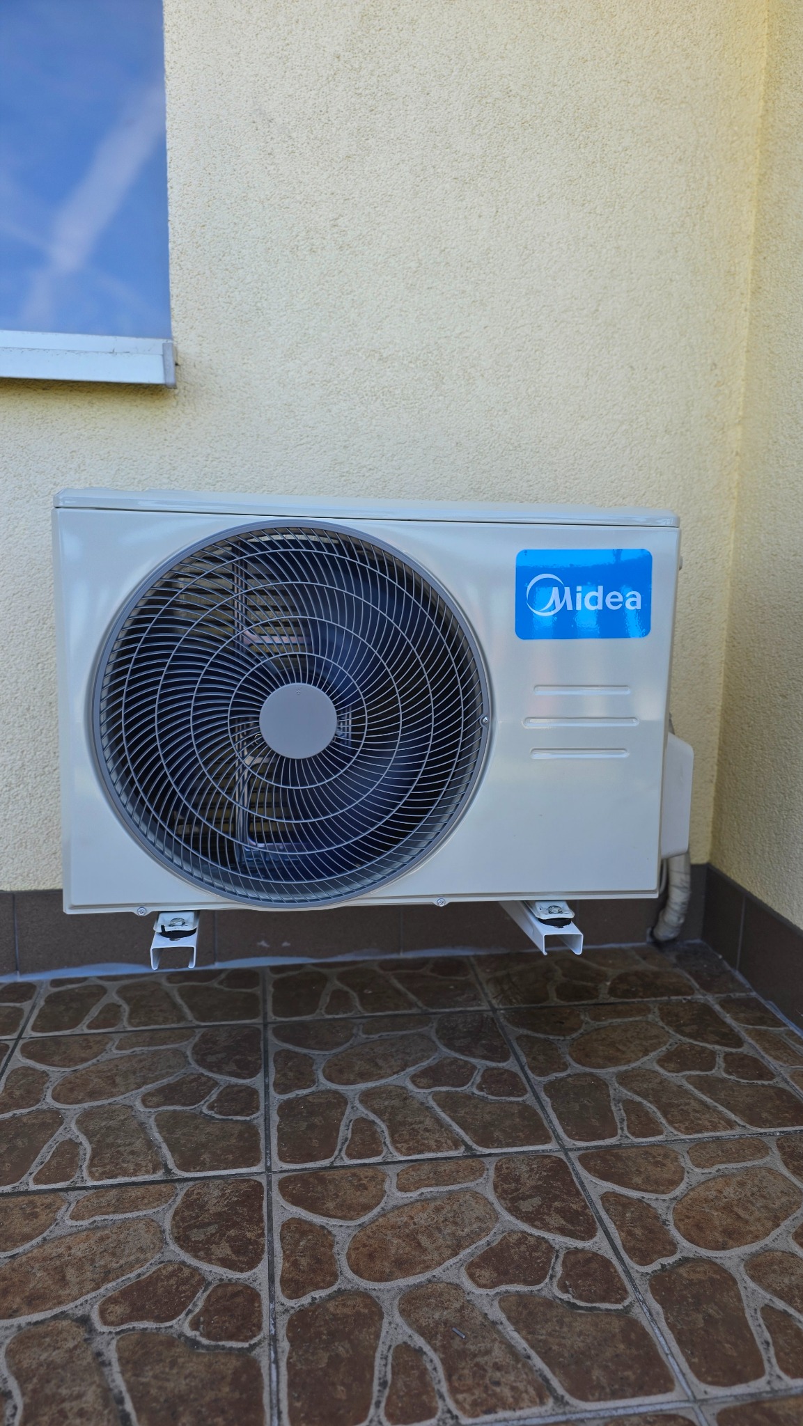 Zewnętrzna jednostka klimatyzacji Midea zamontowana na elewacji budynku z beżowym tynkiem, widoczne brązowe płytki na posadzce.