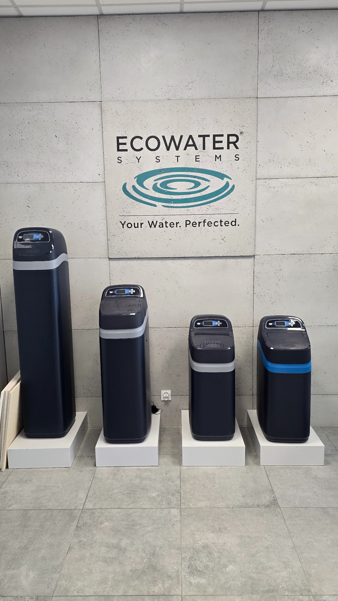 Cztery urządzenia do uzdatniania wody marki Ecowater Systems, różniące się wysokością i pojemnością, ustawione obok siebie na białych podestach na tle szarej ściany z logo firmy.