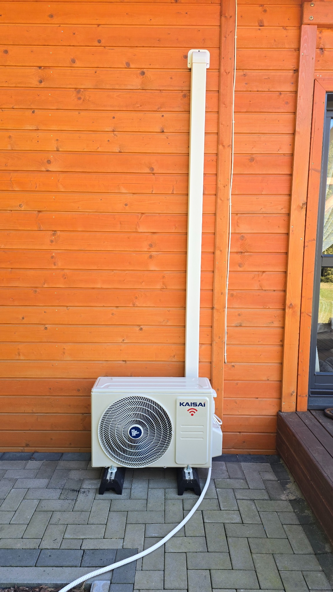 Zewnętrzna jednostka klimatyzacji Kaisai zamontowana na czarnych wspornikach przy drewnianej ścianie domu, z widoczną białą rurą odprowadzającą i kostką brukową.