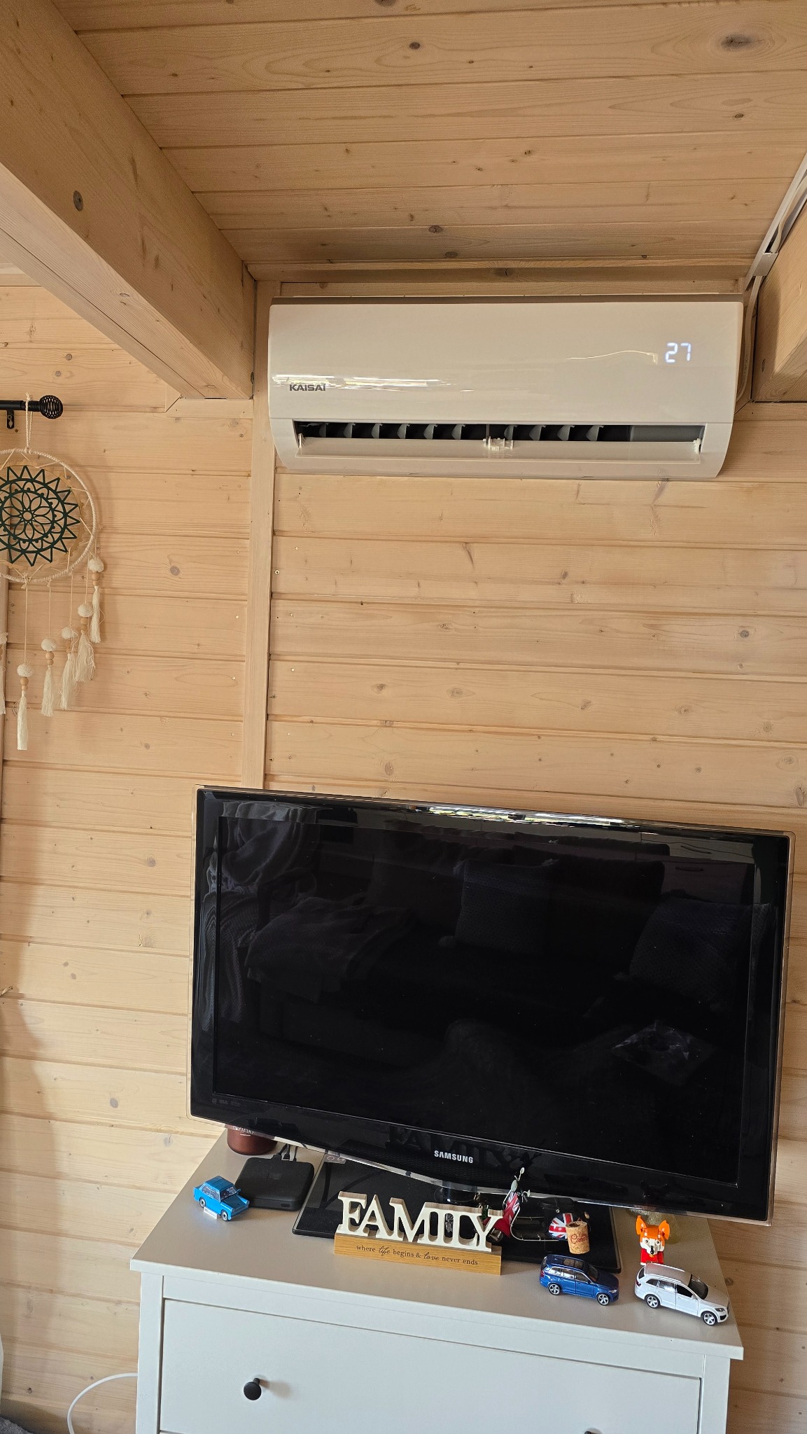 Klimatyzator Kaisai zamontowany na drewnianej ścianie domku, obok łapacza snów i nad telewizorem Samsung, temperatura wyświetlana na urządzeniu to 27 stopni.