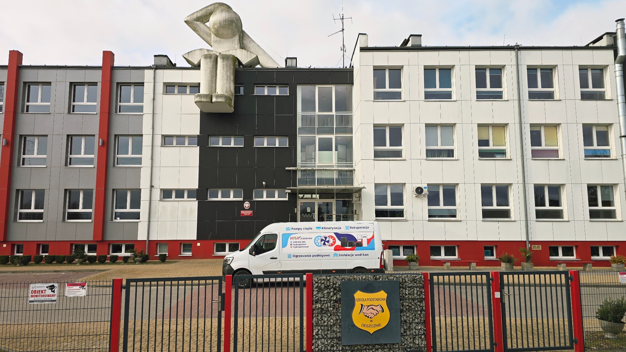 Biały van z reklamą usług (pompy ciepła, klimatyzacja, rekuperacja, ogrzewanie podłogowe) zaparkowany przed budynkiem szkoły podstawowej w Deszcznie.