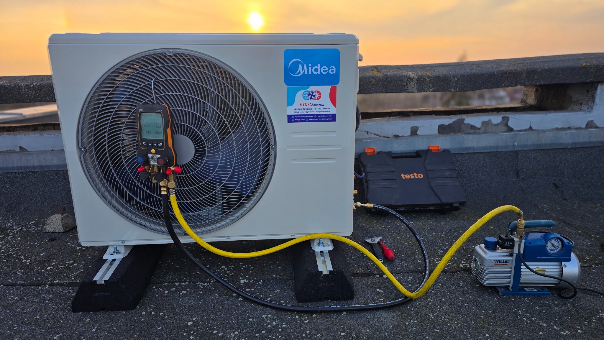 Biała jednostka zewnętrzna klimatyzacji Midea na dachu, podłączona do pompy próżniowej i manometru cyfrowego Testo, z zachodem słońca w tle. Widoczne narzędzia i akcesoria.