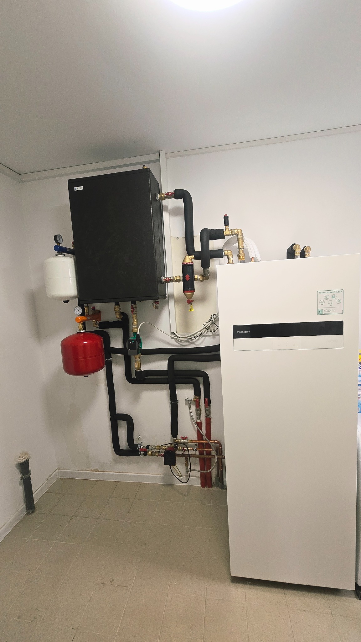 Zestaw instalacji grzewczej z czarnymi rurami, czerwonym zbiornikiem i białym panelem Panasonic Aquarea Smart Cloud na tle białej ściany i beżowej podłogi.