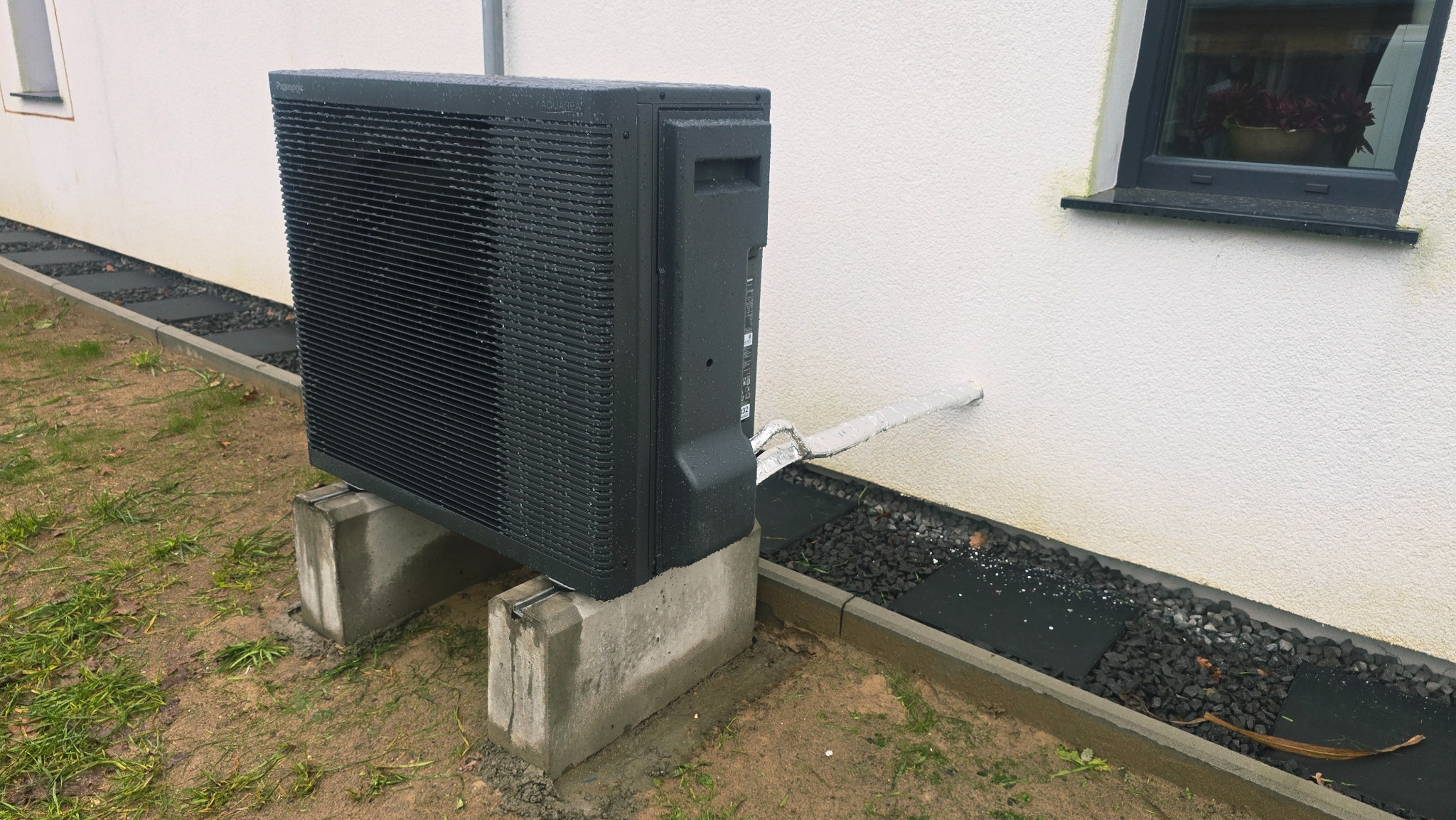 Czarna pompa ciepła Aquarea Panasonic na betonowych podstawach, zamontowana przy białej ścianie domu z oknem. Widoczne rury odprowadzające.