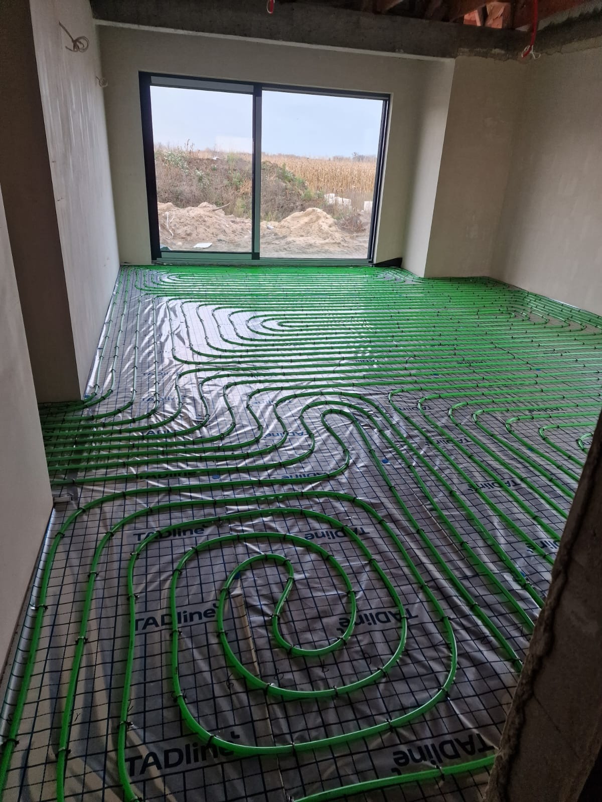 Zielone rury ogrzewania podłogowego ułożone na folii z siatką w surowym wnętrzu z oknem na zewnątrz. Widoczne elementy mocujące rury.