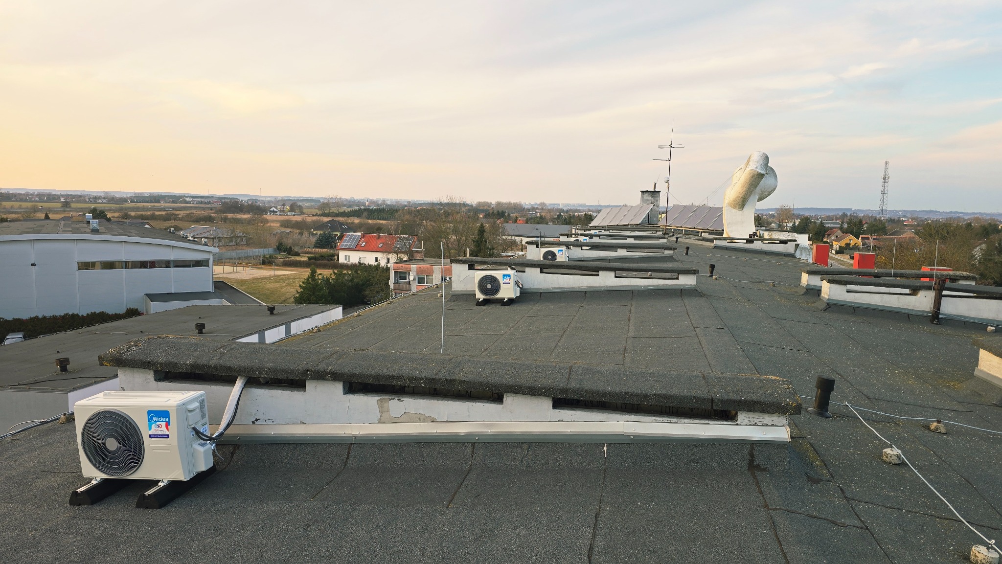 Kaskada klimatyzatorów Midea na płaskim dachu budynku, widok z góry na tle zabudowań i zachodzącego słońca. Instalacja klimatyzacji na dachu.