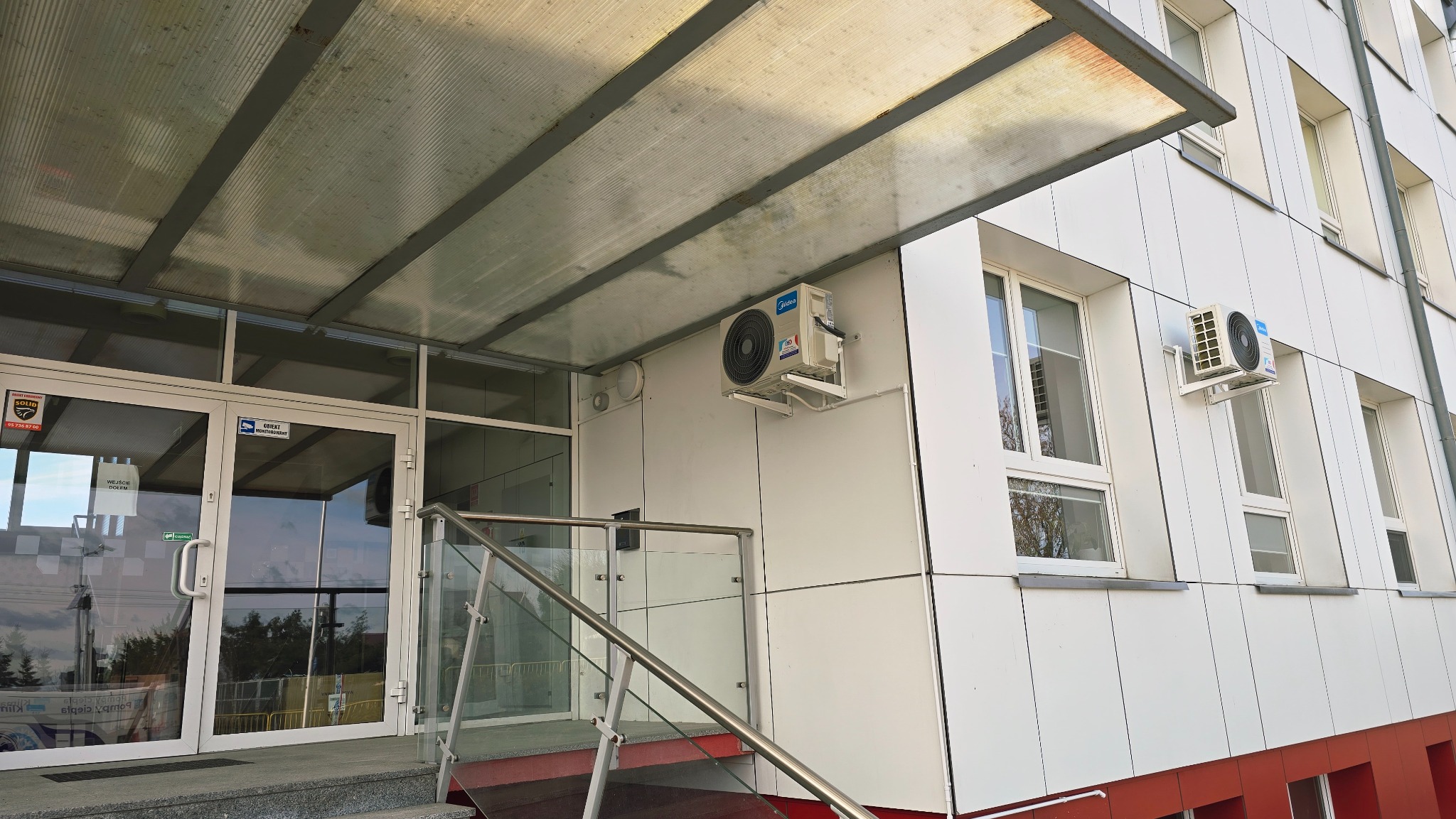 Biała fasada budynku z dwoma zamontowanymi klimatyzatorami Midea. Wejście z przeszklonymi drzwiami i zadaszeniem. Widok z dołu, lekko z prawej strony.