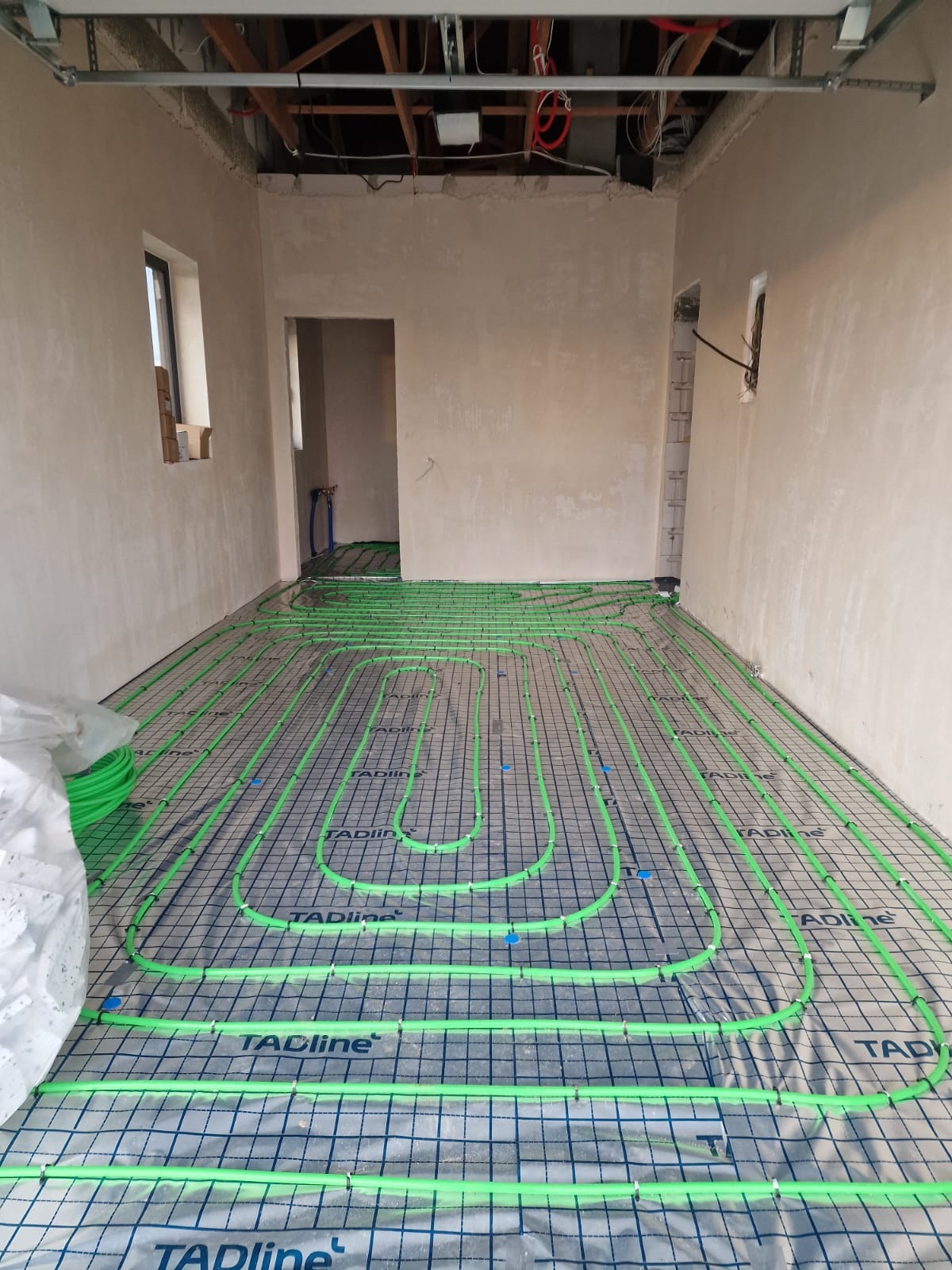 Wnętrze w trakcie montażu ogrzewania podłogowego: zielone rurki układane na siatce, surowy tynk na ścianach, widoczne instalacje pod sufitem. Etap przed wylaniem posadzki.