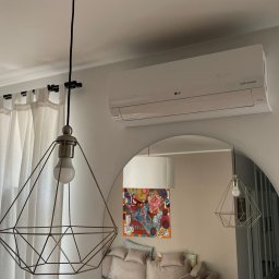 GRUPA ENERGYPRO SP. Z O.O. - Biała jednostka klimatyzacji LG zamontowana na ścianie nad lustrem łukowym, z widoczną lampą wiszącą o geometrycznym kształcie i zasłonami w tle.