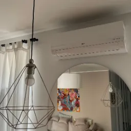Biała jednostka klimatyzacji LG zamontowana na ścianie nad lustrem łukowym, z widoczną lampą wiszącą o geometrycznym kształcie i zasłonami w tle.