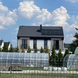 GRUPA ENERGYPRO SP. Z O.O. - Nowoczesny dom jednorodzinny z panelami fotowoltaicznymi na dachu, widok zza ogrodzenia i folii tunelowych, błękitne niebo z obłokami w tle.