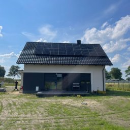 GRUPA ENERGYPRO SP. Z O.O. - Dom jednorodzinny z panelami słonecznymi na dachu, widok z zewnątrz, na trawniku leży zwinięty wąż, w tle osoba przechodzi obok materiałów budowlanych.