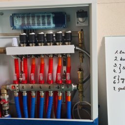 GRUPA ENERGYPRO SP. Z O.O. - Rozdzielacz ogrzewania podłogowego w białej szafce, z rurami w kolorach czerwonym i niebieskim, widoczny obok biała tablica z odręcznie napisaną listą pomieszczeń.