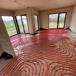 GRUPA ENERGYPRO SP. Z O.O. - Pomieszczenie w stanie surowym z dużymi oknami, w którym ułożono pomarańczowe rury ogrzewania podłogowego na folii izolacyjnej i siatce.