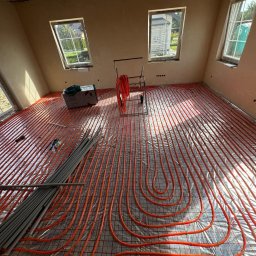 GRUPA ENERGYPRO SP. Z O.O. - Pomieszczenie w trakcie instalacji ogrzewania podłogowego: czerwone rurki ułożone na folii izolacyjnej z siatką, widoczne okna i przygotowane rury do podłączenia.