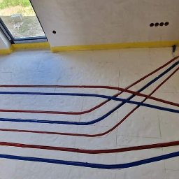 GRUPA ENERGYPRO SP. Z O.O. - Rozłożone rury ogrzewania podłogowego w kolorze niebieskim i czerwonym na izolacji, widoczna rozdzielnia z podłączonymi rurami, zabezpieczone ściany taśmą malarską.