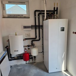 GRUPA ENERGYPRO SP. Z O.O. - Wnętrze kotłowni z zainstalowaną białą pompą ciepła Midea, zbiornikiem Ferroli, czerwonym naczyniem przeponowym i czarnymi rurami instalacji grzewczej.