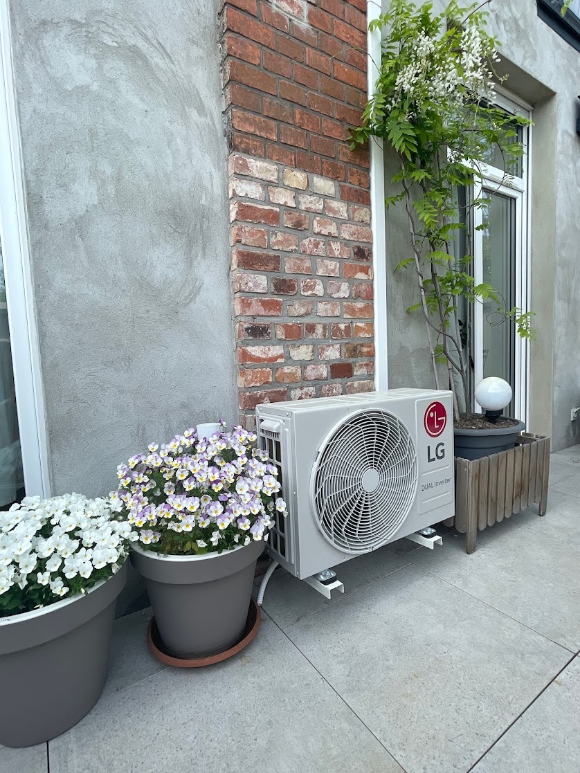 Zewnętrzna jednostka klimatyzacji LG Dual Inverter zamontowana na tarasie, obok donic z białymi i fioletowymi bratkami, z ceglanym narożnikiem budynku w tle i pnącą rośliną przy oknie.
