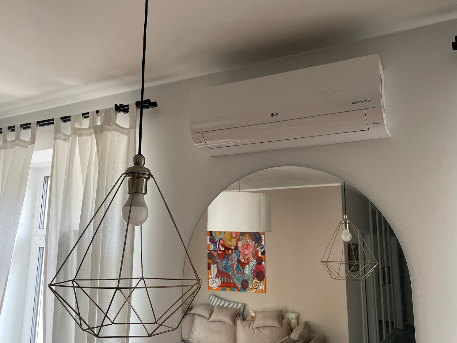 Biała jednostka klimatyzacji LG zamontowana na ścianie nad lustrem łukowym, z widoczną lampą wiszącą o geometrycznym kształcie i zasłonami w tle.