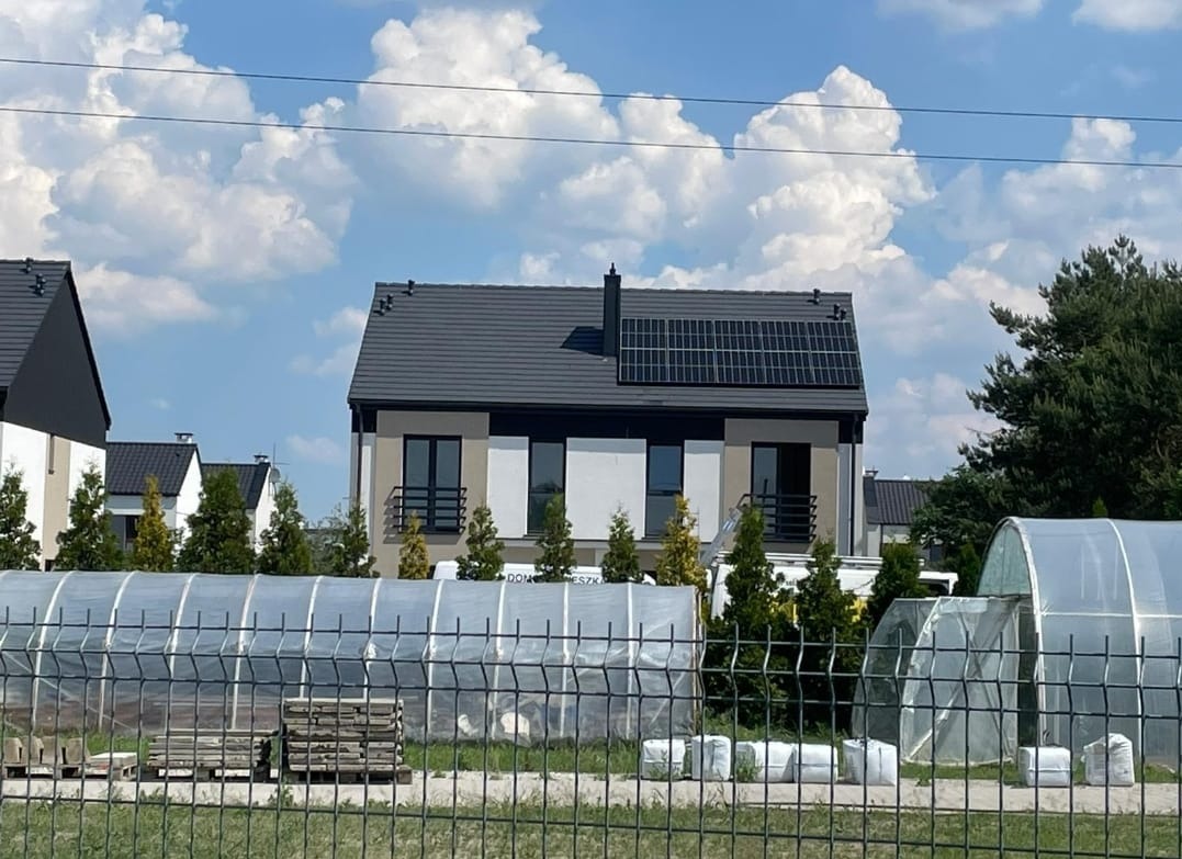 Nowoczesny dom jednorodzinny z panelami fotowoltaicznymi na dachu, widok zza ogrodzenia i folii tunelowych, błękitne niebo z obłokami w tle.