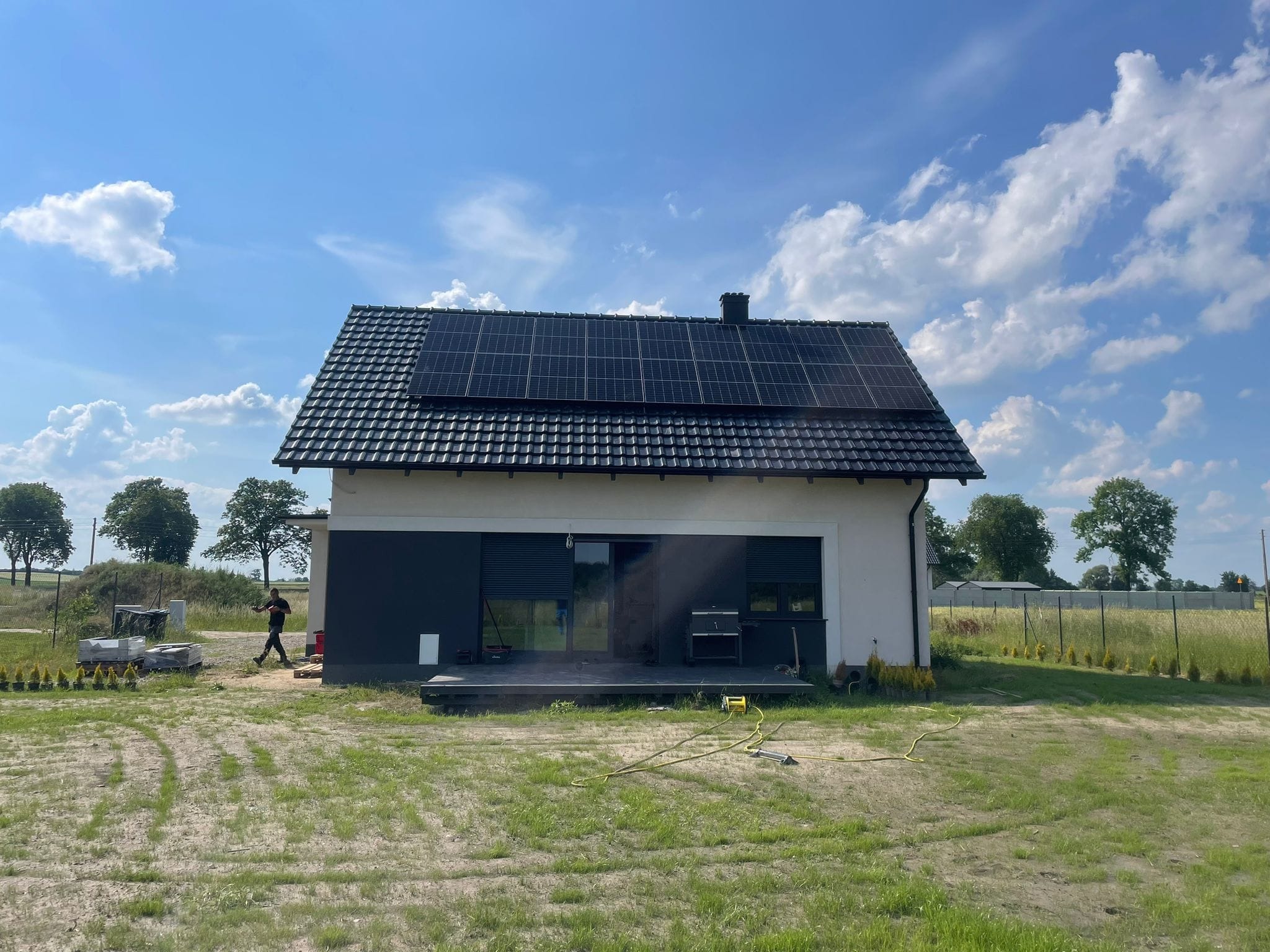 Dom jednorodzinny z panelami słonecznymi na dachu, widok z zewnątrz, na trawniku leży zwinięty wąż, w tle osoba przechodzi obok materiałów budowlanych.