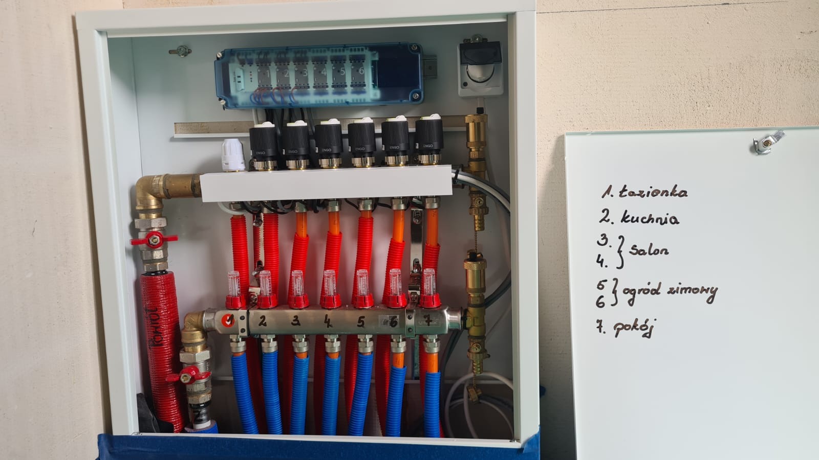 Rozdzielacz ogrzewania podłogowego w białej szafce, z rurami w kolorach czerwonym i niebieskim, widoczny obok biała tablica z odręcznie napisaną listą pomieszczeń.