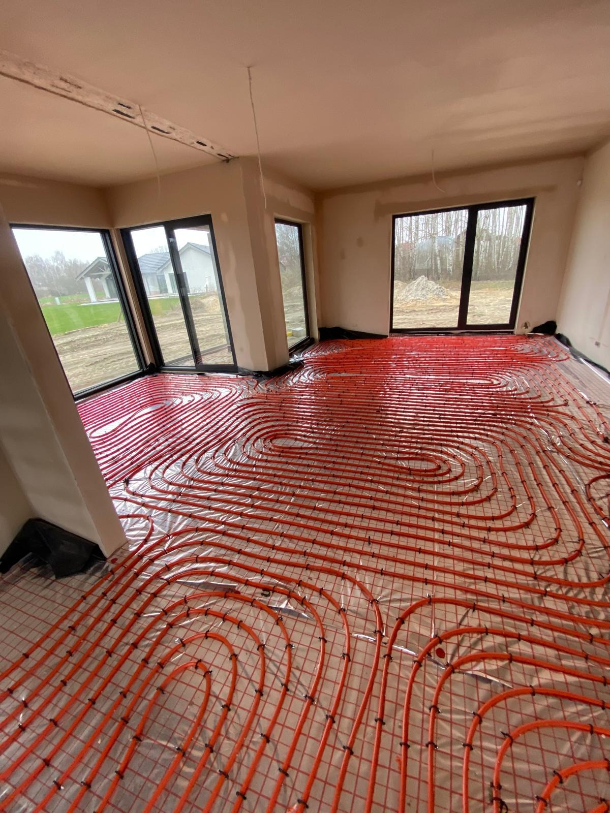 Pomieszczenie w stanie surowym z dużymi oknami, w którym ułożono pomarańczowe rury ogrzewania podłogowego na folii izolacyjnej i siatce.