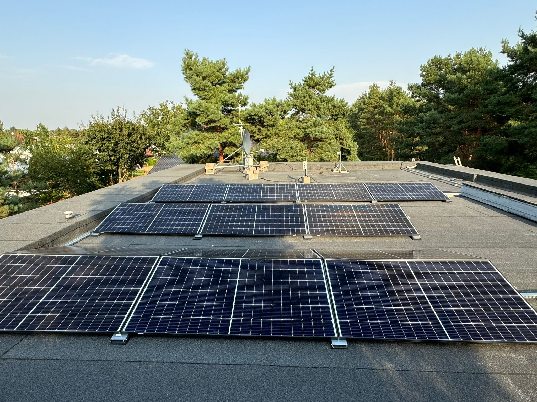Panele słoneczne zamontowane na płaskim dachu pokrytym papą, z widocznym systemem mocowań i anteną satelitarną w tle, otoczone zielenią drzew.