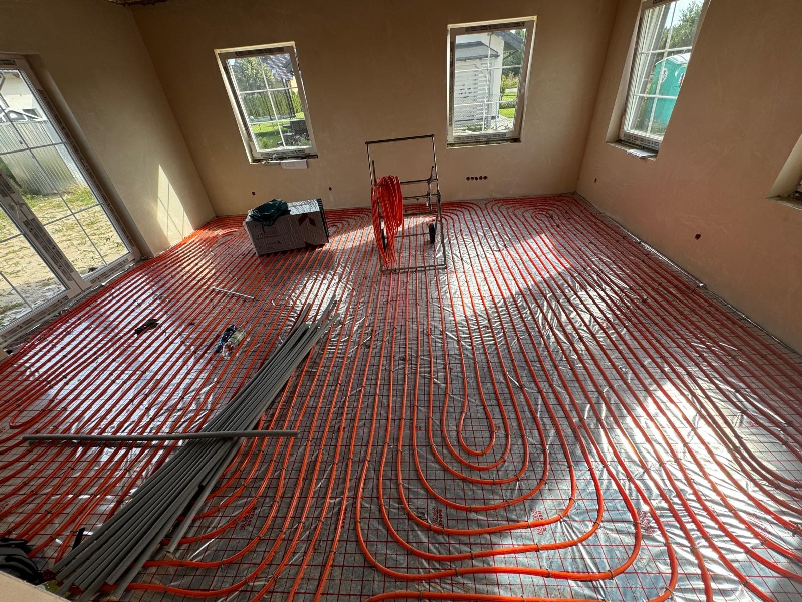 Pomieszczenie w trakcie instalacji ogrzewania podłogowego: czerwone rurki ułożone na folii izolacyjnej z siatką, widoczne okna i przygotowane rury do podłączenia.