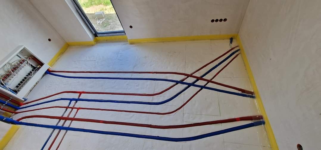 Rozłożone rury ogrzewania podłogowego w kolorze niebieskim i czerwonym na izolacji, widoczna rozdzielnia z podłączonymi rurami, zabezpieczone ściany taśmą malarską.