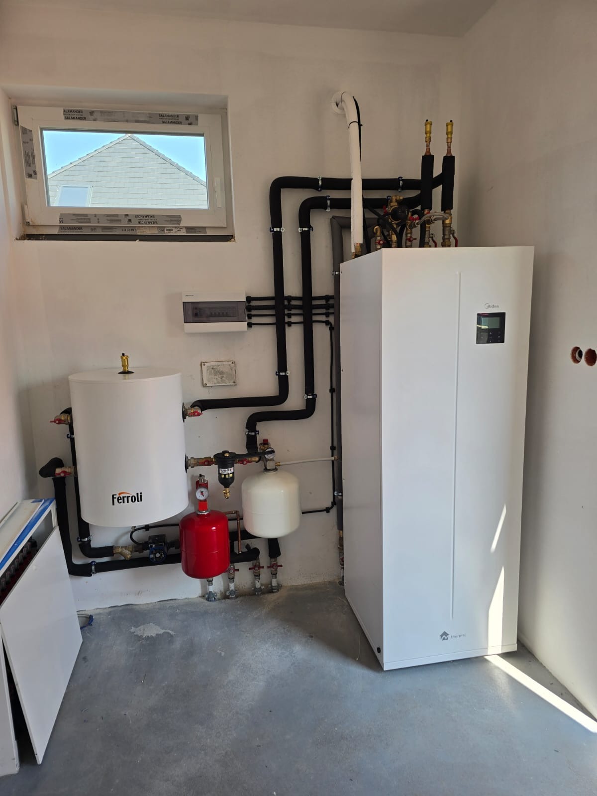 Wnętrze kotłowni z zainstalowaną białą pompą ciepła Midea, zbiornikiem Ferroli, czerwonym naczyniem przeponowym i czarnymi rurami instalacji grzewczej.