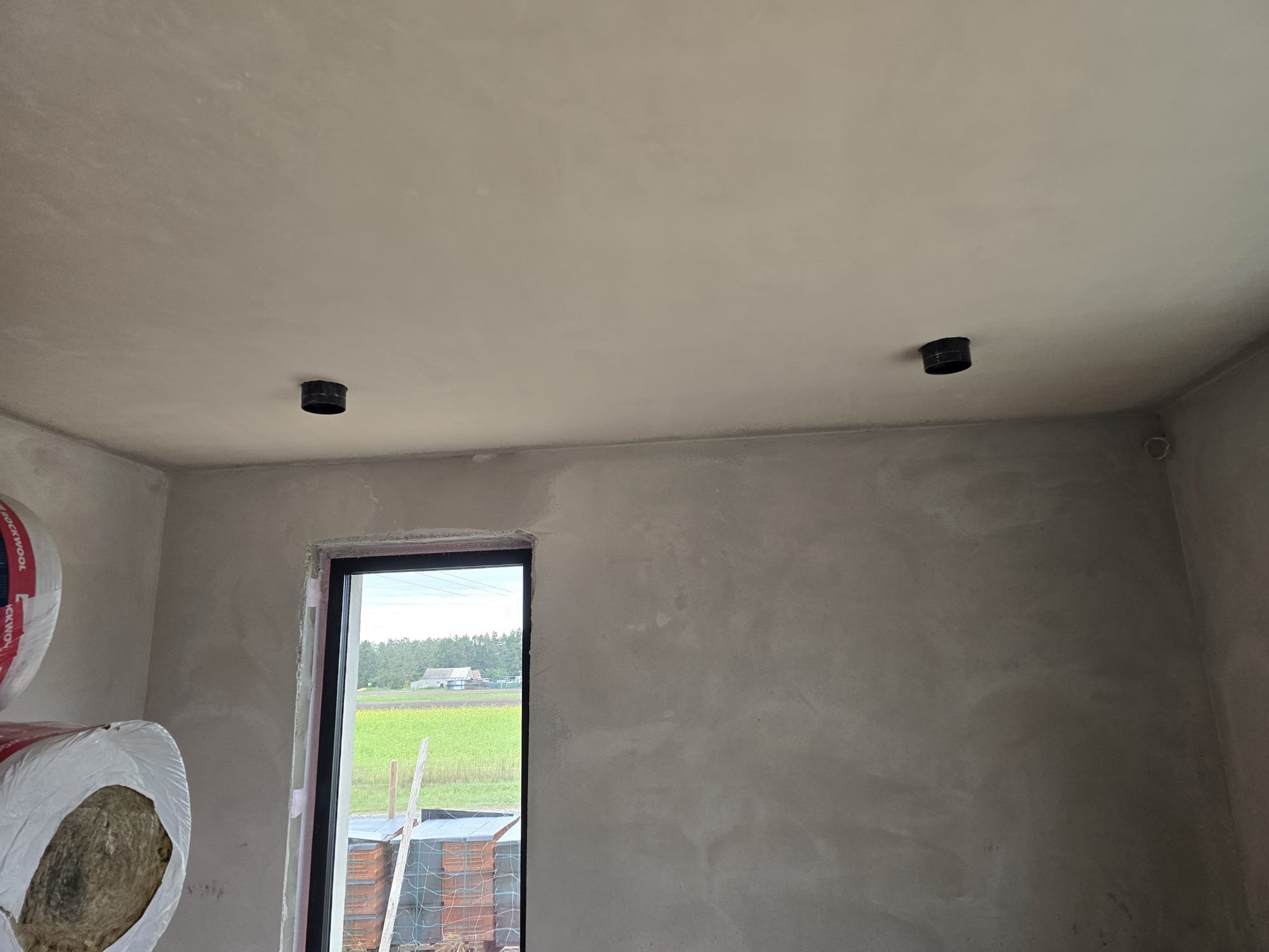 Surowy stan wnętrza w trakcie prac wykończeniowych: ściany pokryte szarym tynkiem, widoczne otwory na instalację wentylacyjną w suficie, rolka wełny mineralnej w rogu, okno z widokiem na pole...