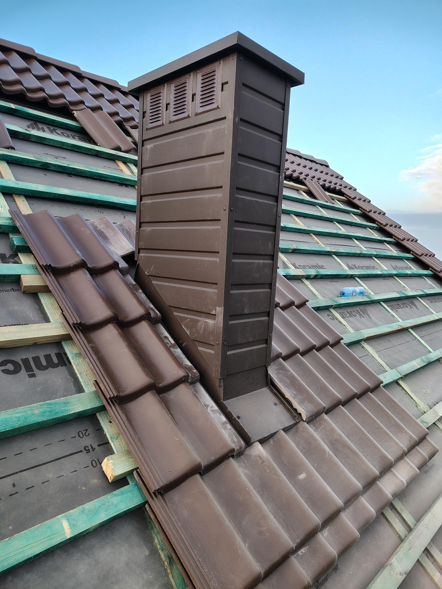 Układanie brązowej dachówki ceramicznej wokół komina wentylacyjnego na dachu z zielonymi łatami i membraną. Widok z góry, częściowo ukończone krycie.