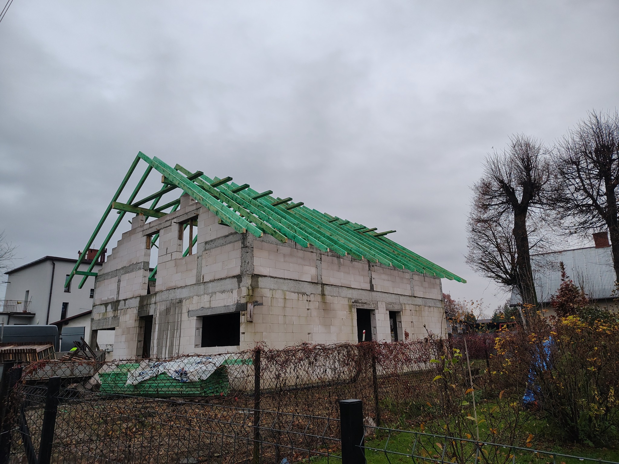 Surowy stan domu z białego pustaka, z częściowo zamontowaną, zieloną konstrukcją dachu. Widoczne materiały budowlane i jesienne otoczenie.