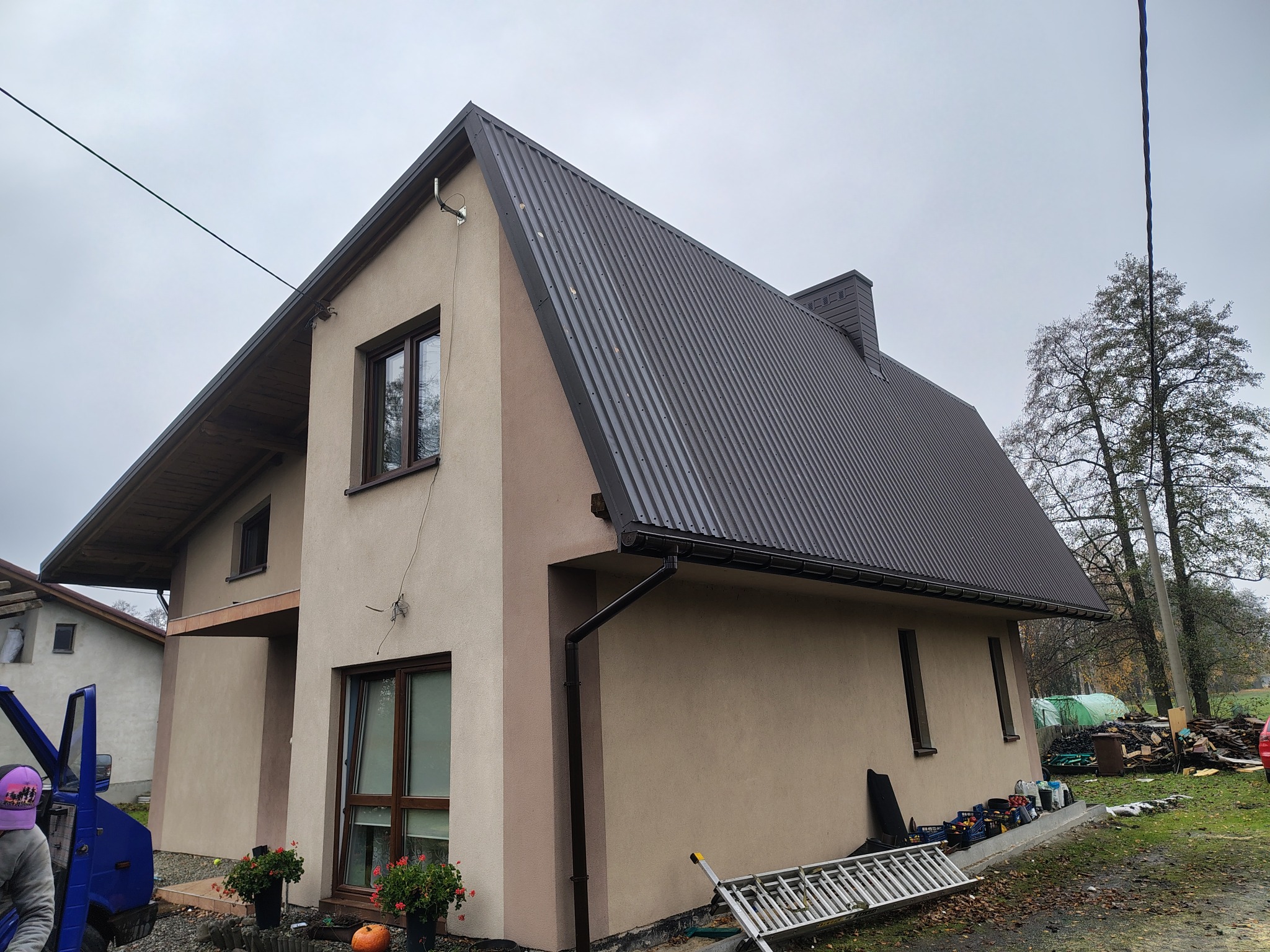 Gotowy dom z dwuspadowym dachem z blachy trapezowej w kolorze grafitowym, z rynnami i oknami. Na pierwszym planie drabina i donice z kwiatami.