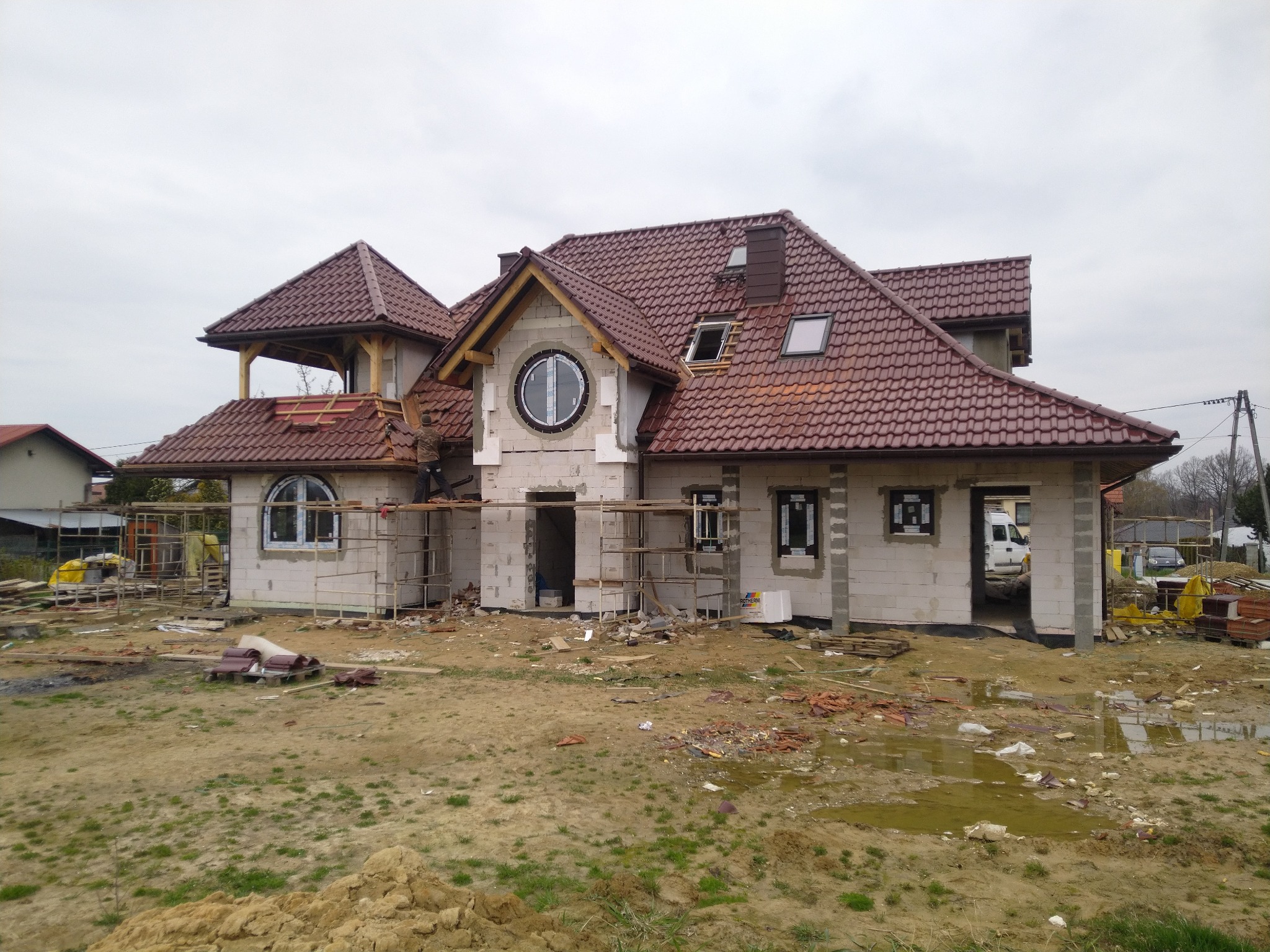 Niedokończony dom murowany z czerwonym dachem i okrągłym oknem, widoczne rusztowania i materiały budowlane na zewnątrz