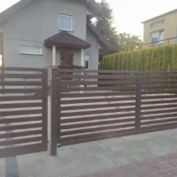 Nowoczesna, pozioma brama przesuwna i furtka w kolorze brązowym, z widocznym domem jednorodzinnym w tle i kostką brukową na podjeździe.