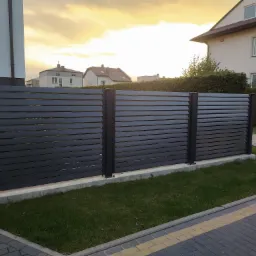 Nowoczesne, poziome ogrodzenie z ciemnoszarych paneli na betonowej podmurówce, widok z poziomu chodnika, z domami w tle i zachodzącym słońcem.