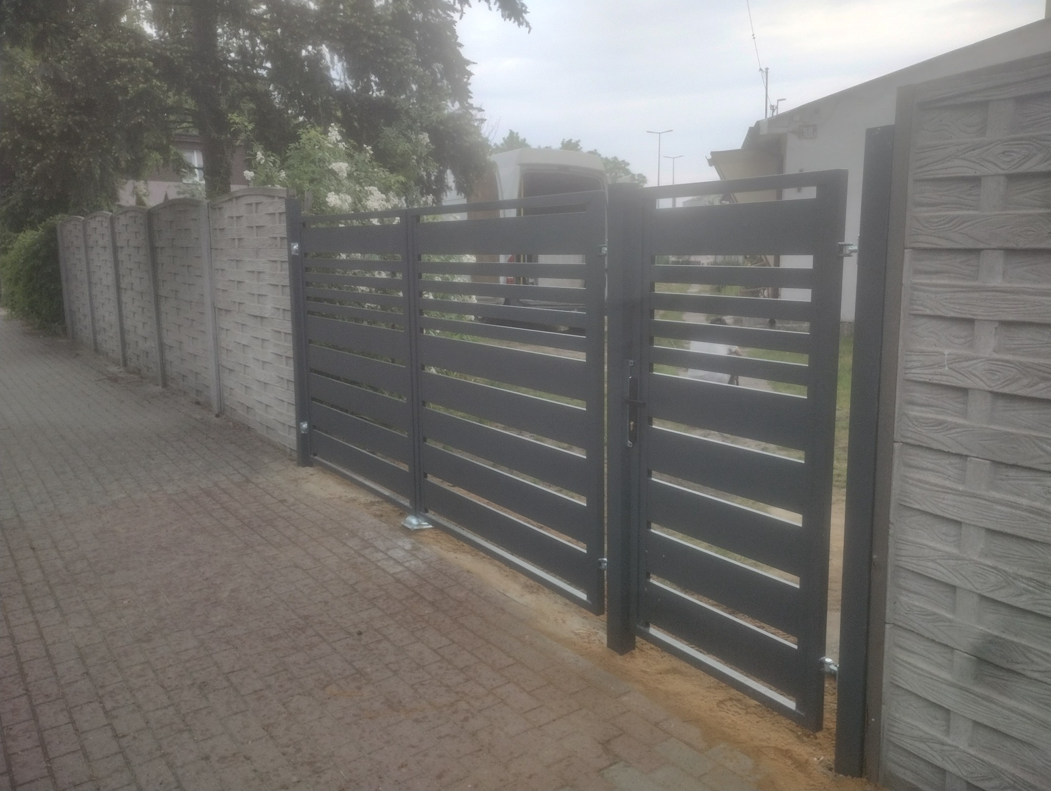 Nowoczesna, pozioma brama wjazdowa w kolorze grafitowym, zamontowana obok betonowego ogrodzenia o wzorze imitującym drewno, na tle brukowanej nawierzchni.