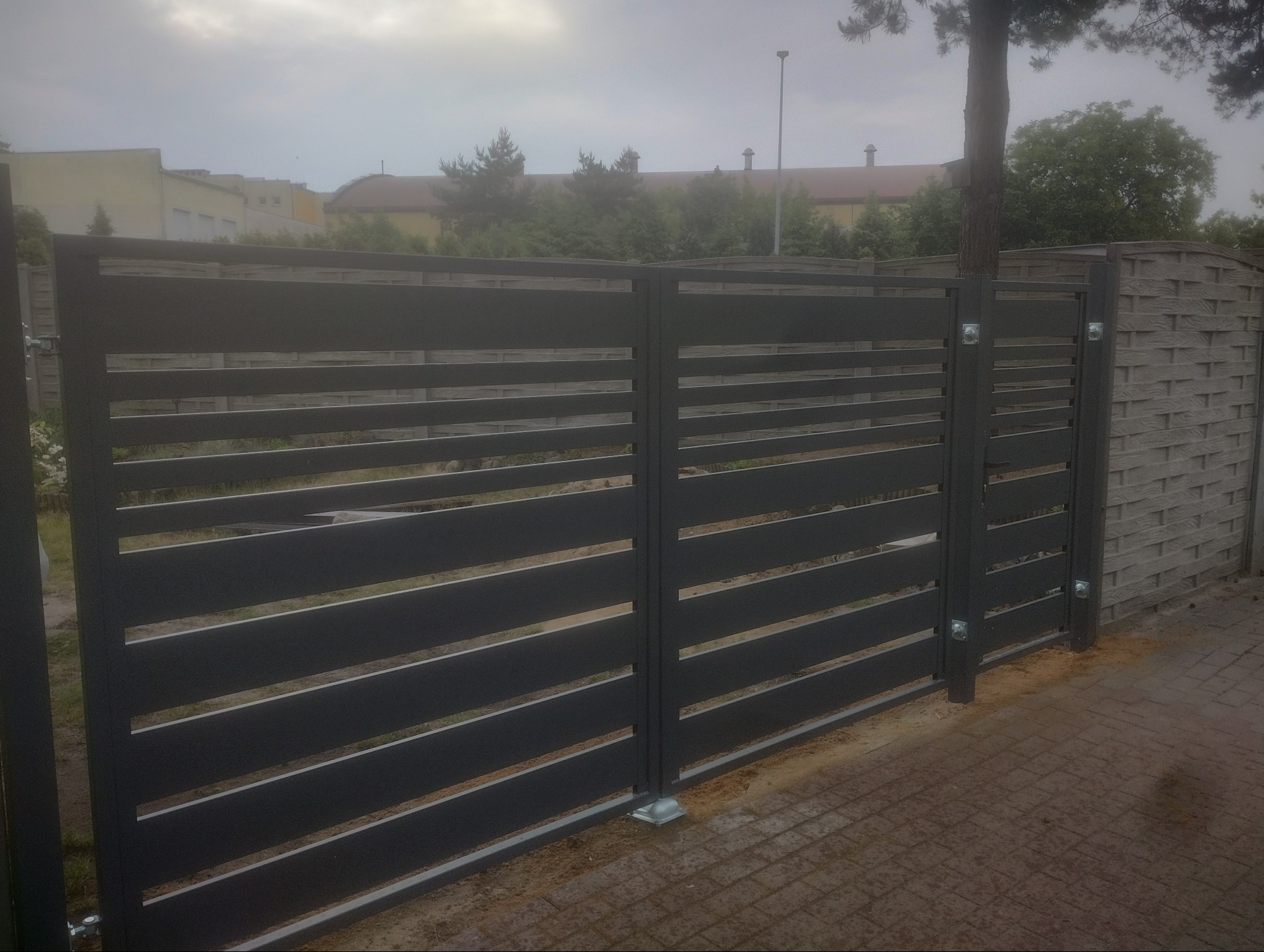 Nowoczesna, pozioma, szara brama wjazdowa z metalowymi przęsłami, widoczna z zewnątrz posesji, obok ogrodzenie betonowe imitujące plecionkę, na tle zabudowań i drzew w pochmurny dzień.