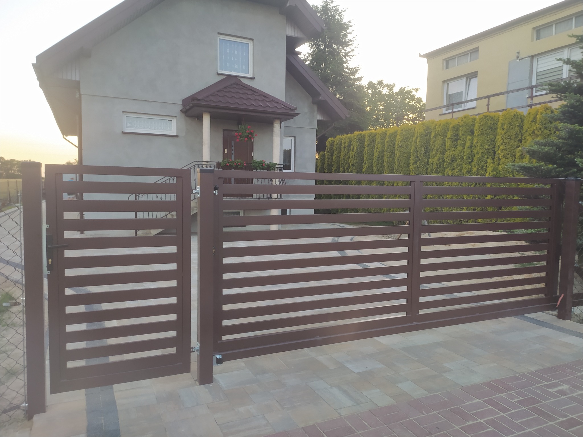 Nowoczesna, pozioma brama przesuwna i furtka w kolorze brązowym, z widocznym domem jednorodzinnym w tle i kostką brukową na podjeździe.