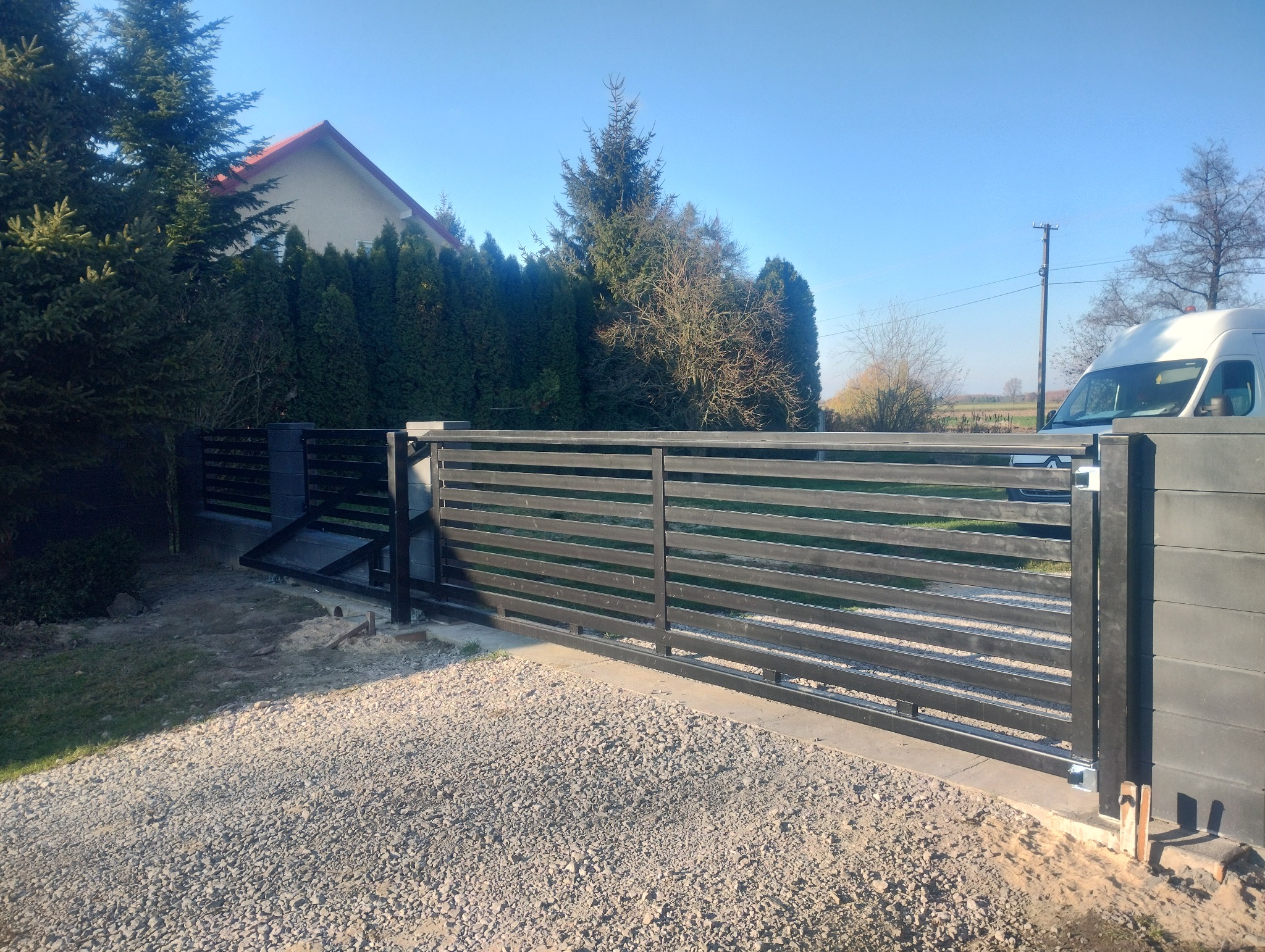 Nowoczesna brama wjazdowa przesuwna z poziomymi panelami, zamontowana na posesji z zielonymi tujami i widocznym fragmentem domu oraz białym samochodem dostawczym w tle.