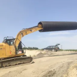 Żółta koparka CAT z czarną rurą drenażową w chwytaku na placu budowy, w tle czarny samochód kombi z otwartym bagażnikiem i błękitne niebo.