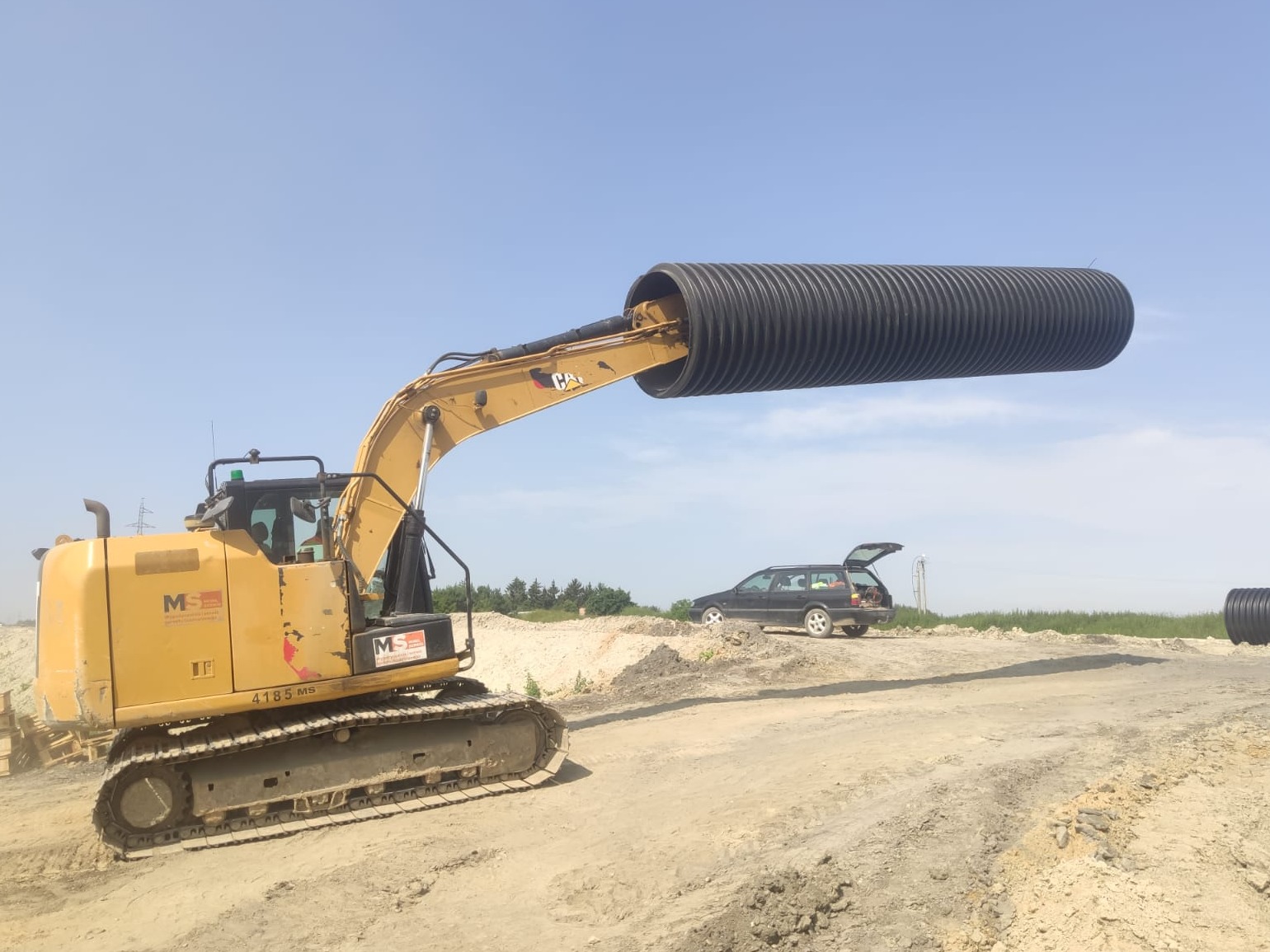 Żółta koparka CAT z czarną rurą drenażową w chwytaku na placu budowy, w tle czarny samochód kombi z otwartym bagażnikiem i błękitne niebo.