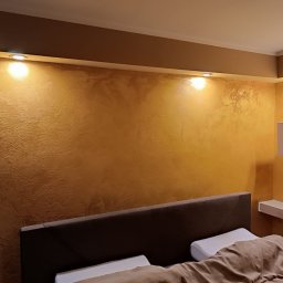 Wall Crafter Remonty ROBERT SIERSZEŃ - Ściana w sypialni z dekoracyjną, złoto-brązową fakturą, oświetlona punktowymi lampami sufitowymi, widoczny fragment łóżka z wezgłowiem i biała półka narożna z dekoracjami.
