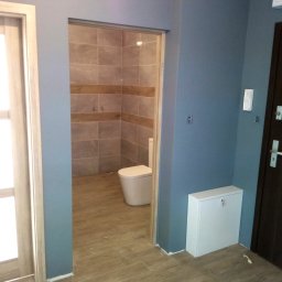 Wall Crafter Remonty ROBERT SIERSZEŃ - Widok na wykończoną łazienkę z toaletą, beżowe płytki na ścianach i podłodze, widoczne z korytarza z niebieskimi ścianami i brązowymi drzwiami wejściowymi.