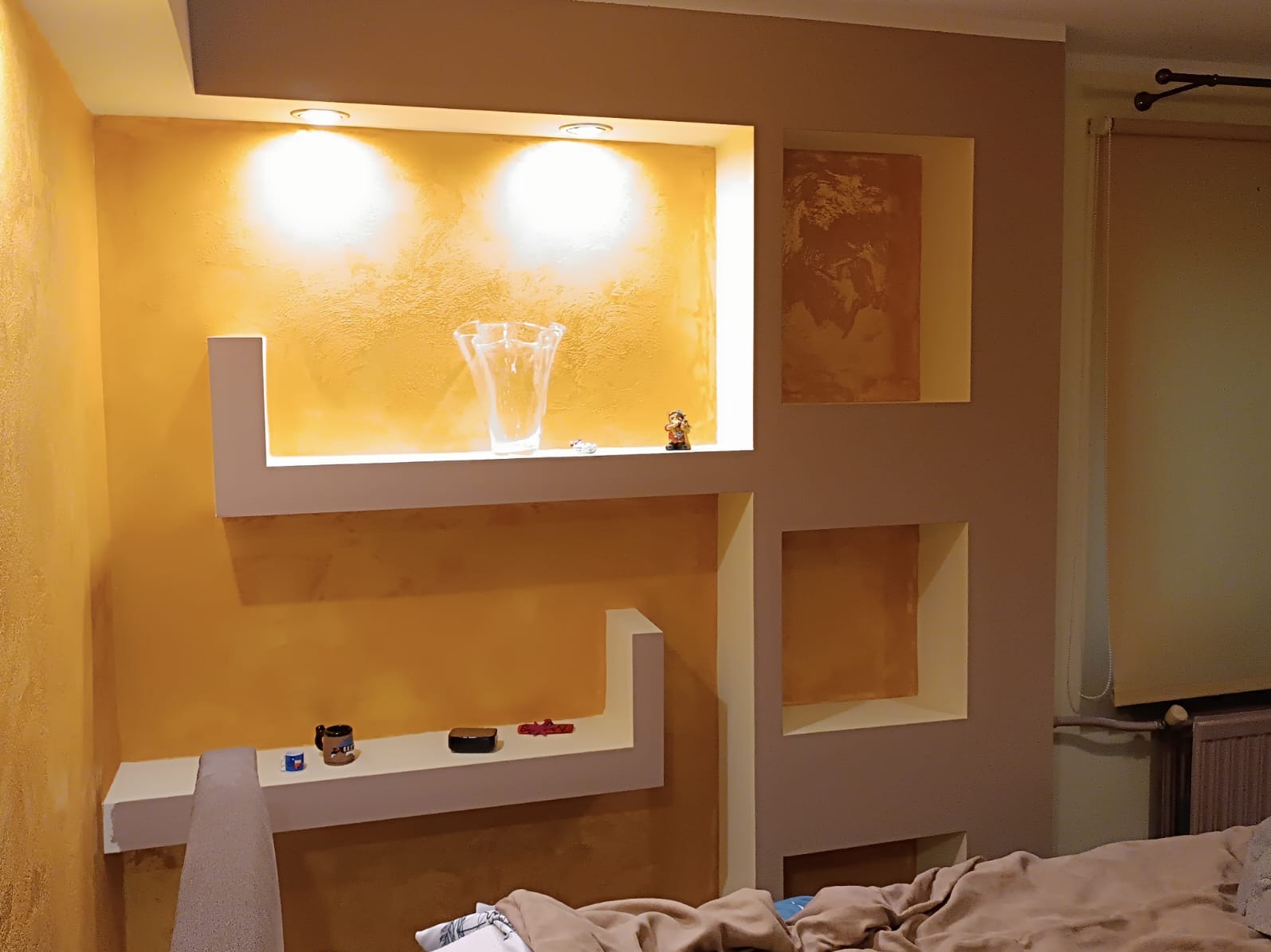 Ściana z wnękami i półkami, pomalowana na żółto i beżowo, z dekoracjami i oświetleniem punktowym, widoczne elementy wystroju wnętrza.