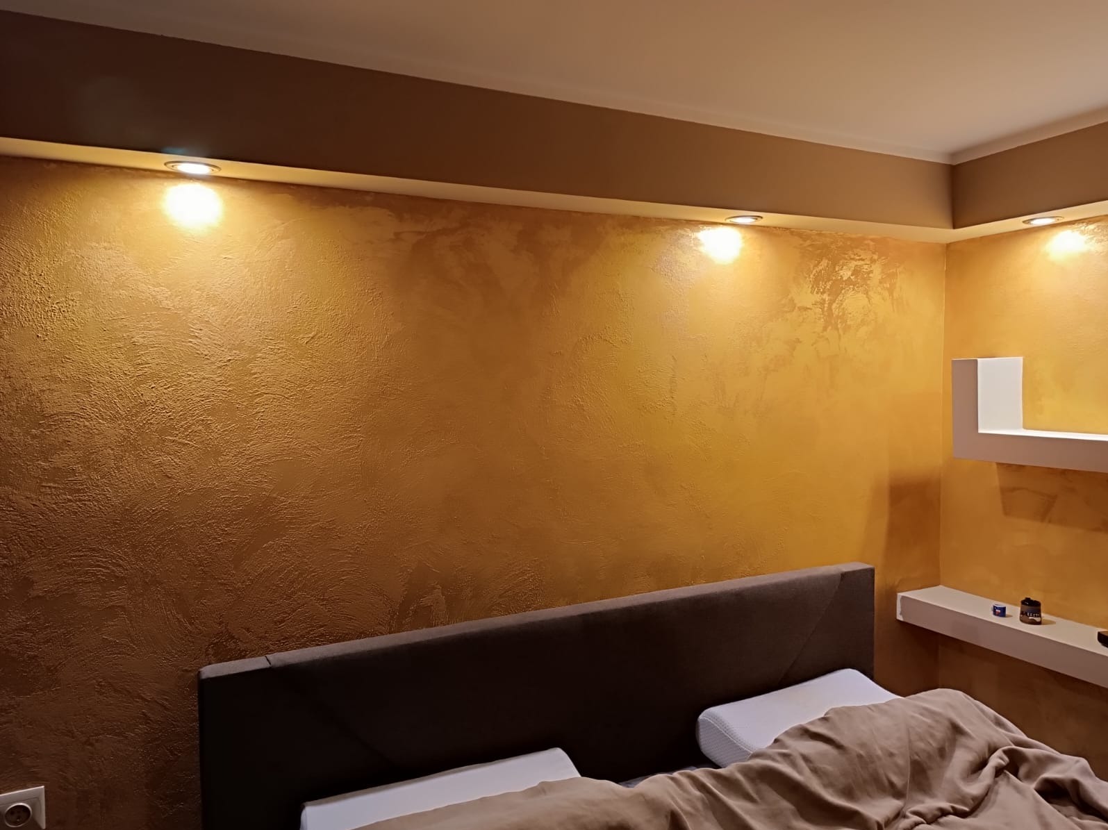 Ściana w sypialni z dekoracyjną, złoto-brązową fakturą, oświetlona punktowymi lampami sufitowymi, widoczny fragment łóżka z wezgłowiem i biała półka narożna z dekoracjami.
